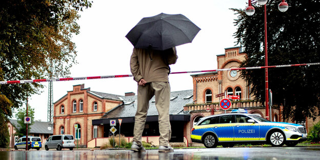 Person, von hinten zu sehen, Kopf durch einen Regschirm verdeckt, steht an einem Polizeiabsperrband