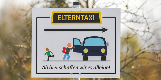 Ein Schild mit der Aufschrift "Elterntaxi Ab hier schaffen wir es alleine!" steht am Parkplatz eines Schulgeländes.