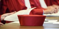 Ein roter Richterhut liegt auf einem Tisch, im Hintergrund sieht man Richter in roten Roben