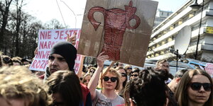 Auf einer Demonstration hält eine blonde Frau mit Sinnenbrille ein Plakat hoch, auf dem ein Uterus den Mittelfinger zeigt.