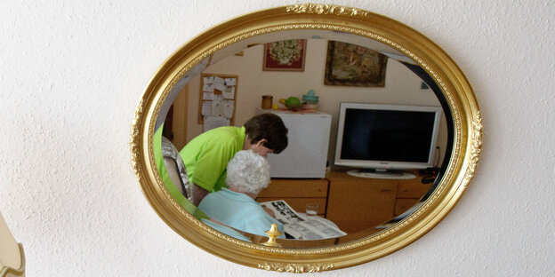 In einem Spiegel ist zu sehen, wie sich eine stehende Pflegekraft und eine sitzende, ältere Person über ein Fotoalbum beugen