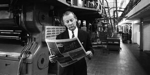 Ein Mann steht vor einer großer Druckmaschine und liest eine Zeitung (Schwarz-Weiß-Foto)
