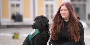 Eine junge Frau sitzt neben ihrem Assistenzhund, einem schwarzen Labrador