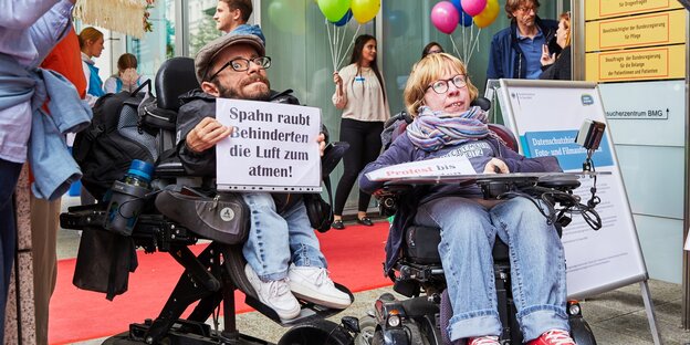 Ein mann und eine Frau im elektro-Rollstuhl, er hält ein Plakat, dass sich gegen Soahns Gesetzentwurf zur Intensivpflege wendet