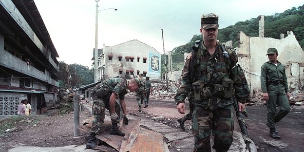 Soldaten gehen auf einer Strasse, im Hintergrund sieht man zerstörte Häuser
