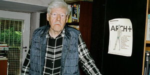 Ein gutaussehender alter Herr im Karohemd steht vor einem Bücherschrank, an dem ein altes Titelbild der arch+ hängt