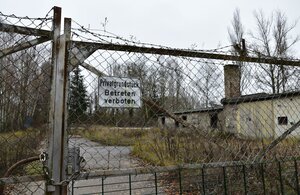 Hinter einem verschlossenen Tor mit Zaungitter und Stacheldraht steht ein altes Gemäuer zwischen Gestrüpp. Ein Schornstein ragt auf und die Fenster sind eingeschlagen. Hinter einem verschlossenen Tor mit Zaungitter und Stacheldraht steht ein altes Gemäuer zwischen Gestrüpp. Ein Schornstein ragt auf und die Fenster sind eingeschlagen.