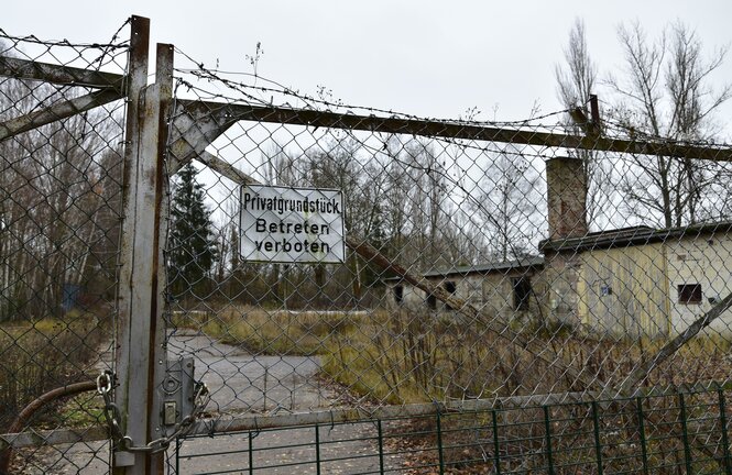 Hinter einem verschlossenen Tor mit Zaungitter und Stacheldraht steht ein altes Gemäuer zwischen Gestrüpp. Ein Schornstein ragt auf und die Fenster sind eingeschlagen.