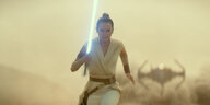 Rey rennt mit ihrem Lichtschwert in der Hand durch die Wüste. Hinter ihr fliegt ein feindliches Kampfschiff