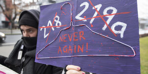 Eine Frau hält ein lilanes Tranparent, auf dem die durchgestrichenen Paragrafen 218 und 219a stehen und ein Kleiderbügel klebt. Dazu die Aufschrift "Never again!"