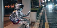 ein als Raumfahrer kostümierter Mensch sitzt auf einer Bank nachts an einer Straße und guckt auf ein Handy mit Handy