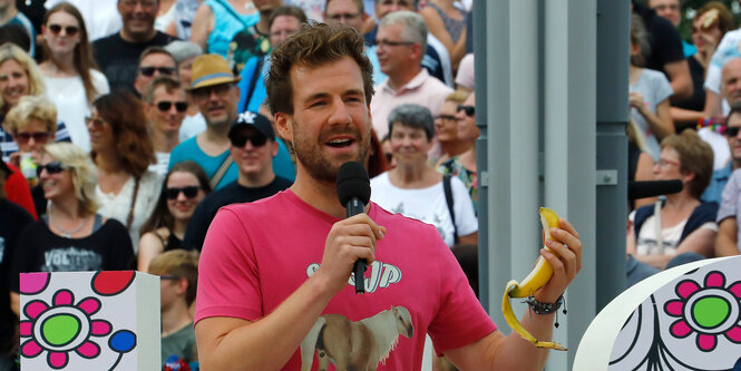 Bärtiger Mann in pinkem Shirt mit Mikrophon und Banane in der Hand