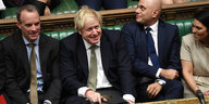 Boris Johnson sitzt zwischen Parteigefährten im Unterhaus und grinst