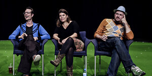 Auf einer grün ausgelegten Bühne sitzen Stefan Kaegi, Helgard Haug und Daniel Wetzel nebeneinander auf Stühlen und lachen.