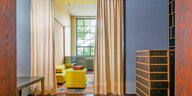 Blick in ein helles Zimmer mit knallgelben Polstermöbeln, hellblauen Regalen und Fensterrahmen
