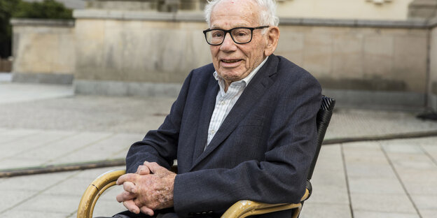 Ein älterer Herr mit Anzug und Brille sitzt auf einem Stuhl auf einem Platz draußen.