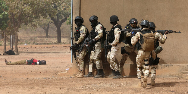 Mehrere Soldaten drängen sich an eine braune Hauswand, im Hintergrund liegen Männer reglos auf dem Boden