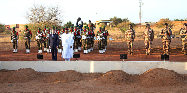 Emmanuel Macron und sein Amtskollege aus Niger Mahamadou Issoufou stehen vor einer niedrigen Steinmauer, davor eine Reihe kleiner Erdhügel. Hinter den Staatschefs stehen Soldaten