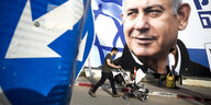 Plakat mit übergroßem Profil von Netanjahu - davor geht ein Mann mit einem Kinderwagen vorbei. Im Vordergrund ein Schild mit einem weißen Pfeil vor blauem Grund