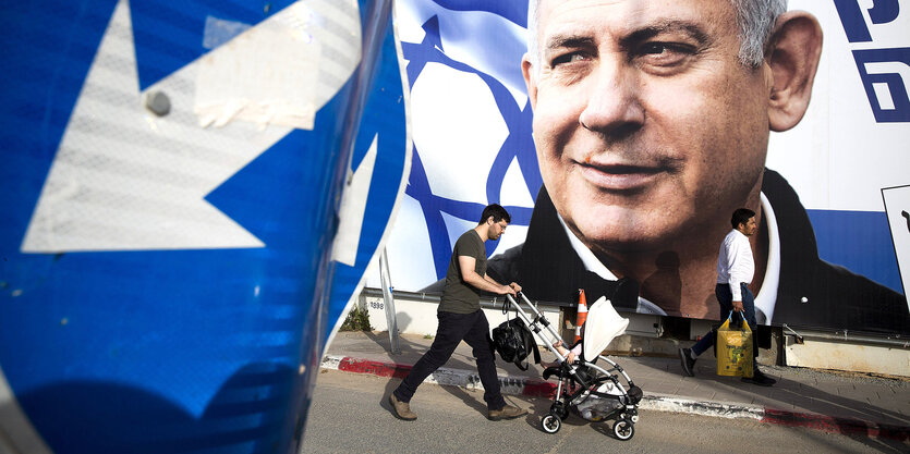 Plakat mit übergroßem Profil von Netanjahu - davor geht ein Mann mit einem Kinderwagen vorbei. Im Vordergrund ein Schild mit einem weißen Pfeil vor blauem Grund