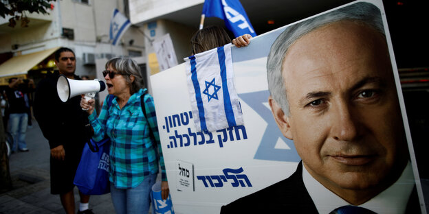 Jemand trägt ein Wahlplakat mit dem Gesicht von Netanjahu. Daneben ruft eine Frau ins Megaphon.