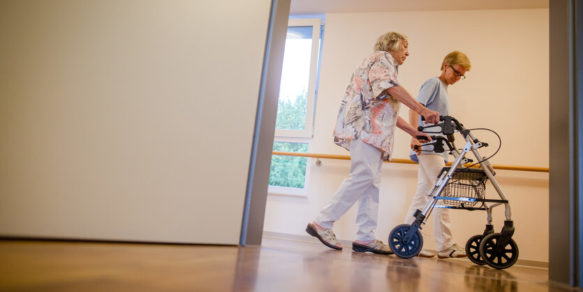 Eine Pflegekraft geht mit einer älterren Frau am Rollator über einen Flur