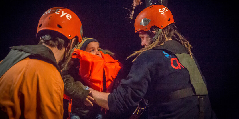 Zwei Männer von der „Alan Kurdi“ mit Helmen heben ein Kleinkind hoch. Es ist Nacht. Das Kind trägt eine Schwimmweste.