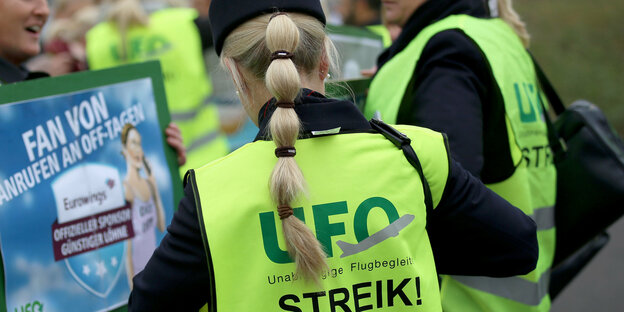 Eine Frau mit Zopf von hinten. Sie trägt eine Warnweste der UFO, auf der "Streik!" steht.