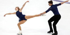 Minerva Hase und Nolan Seegert bei einer Vorführung im Eiskunstlauf. Hase hebt ihr linkes Bein, das Seegert in der Hand hält. Mit dem anderen Bein geht sie in die Knie. Ihr Oberkörper ist nach vorne gebeugt, ihre Arme weit nach Außen gestreckt.