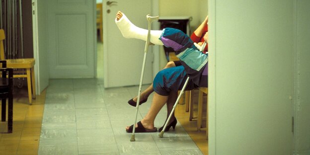 Zwei Personen sitzen im Wartezimmer eines Arztes. Es sind nur die Füße zu sehen, eine Person hat Krücken und einen eingegipstes Bein
