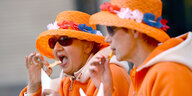 Zwei Damen in orangen Hüten und Jacken essen Pommes mit Mayo