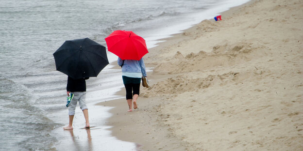 Zwei Menschen mit Regenschirmen laufen am Strand, ein Schirm ist rot, einer ist schwarz