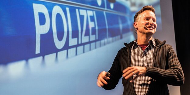 Der Journalist Richard Gutjahr mit einem Ansteckmikro vor einem Beamerbild, darauf steht "Polizei"