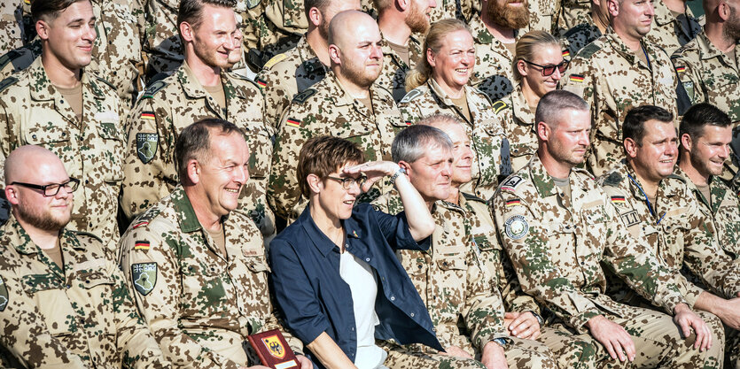 AKK sitzt auf einer Tribüne zwischen einigen Soldaten.