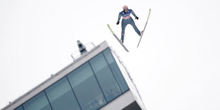 ein Skispringer fliegt auf die Kamera zu. Im Hintergrund die Fensterfront der Schanze