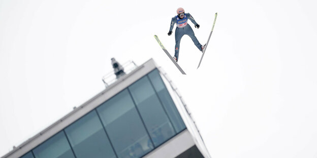 ein Skispringer fliegt auf die Kamera zu. Im Hintergrund die Fensterfront der Schanze
