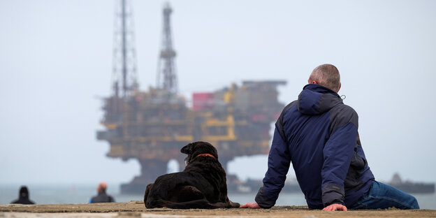 Ein Mann mit Hund sitzt auf einer Sandbank, im Hintergrund auf See eine Bohrinsel