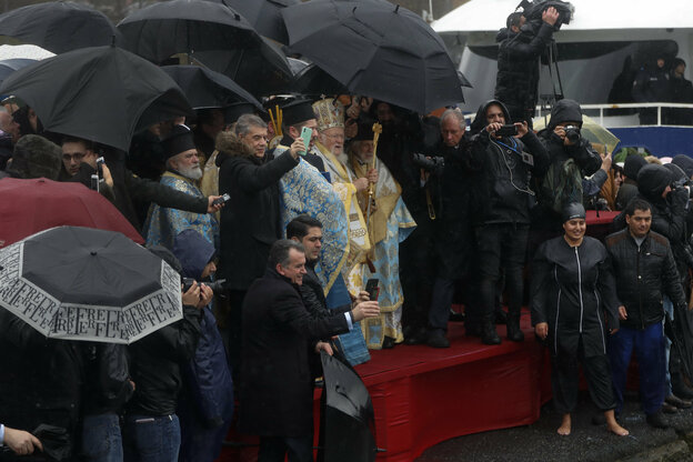 Mann in weißem Gewand mit Krone hält ein Kreuz, um ihn rum viele Menschen mit Regenschirmen