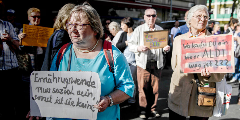 Zwei Frauen protestieren in Berlin gegen die angekündigte Schließung des Discounters Aldi. Auf ihren Schildern steht "Ernährungswende muss sozial sein, sonst ist sie keine" und "Wo kaufst du ein, wenn Aldi weg ist?!"