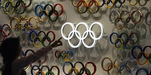 Eine Frai zeigt auf eine Wand auf der die olympischen Ringe ausgestellt sinder Olympischen Spiele und Paralympics 2020