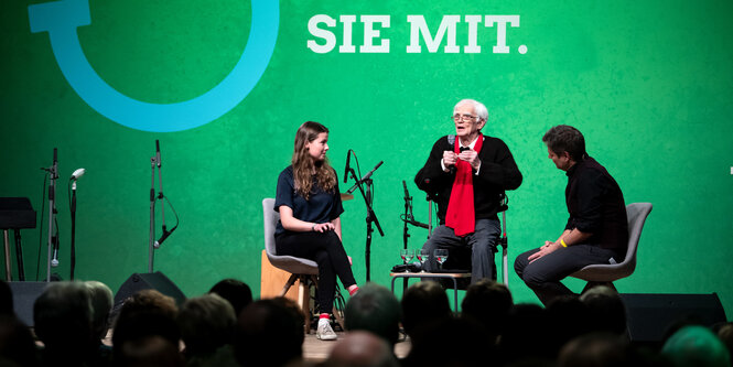 Ein weißhaariger Mann mit einer jungen Frau und einem mittelalten Mann auf einer Bühne