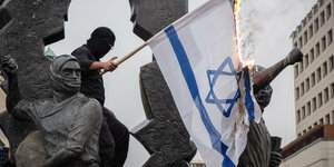 Eine vermummte Person steht auf einem Denkmal. Sie verbrennt eine israelische Flagge.