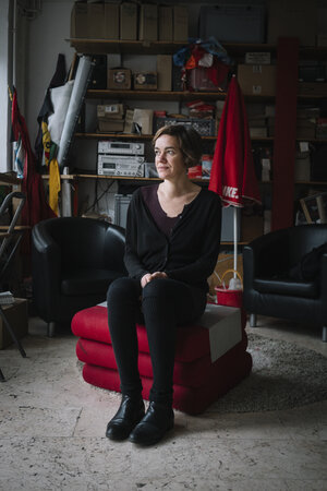 Juliane Nagel sitzt auf in einem Zimmer auf einem Stapel roter Polster. Sie ist dünn und trägt dunkle Klamotten.