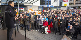 Ein Mann steht auf einer Bühne und spricht in ein Mikrofon. Viele Menschen stehen vor der Bühne und hören zu