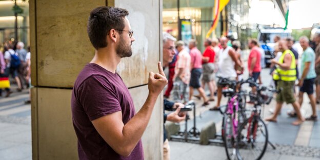 Ein Mann steht hinter einer Gebäudesäule und zeigt einen Stinkefinger in Richtung einer vorbeiziehenden Demo im Hintergrund