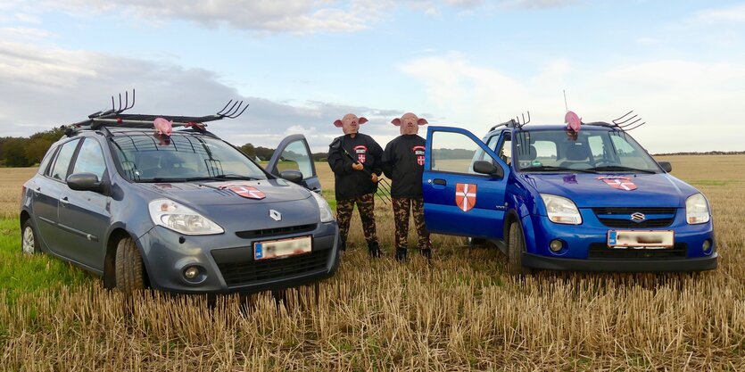 Zwei autos stehen auf einem Feld, dazwischen zwei Männer mit Schweinsmasken und Mistgabeln in der Hand