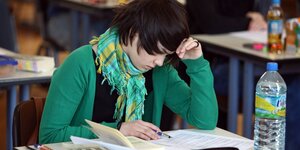 Eine Schülerin sütz ihren Kopf in die Hand, auf dem Tisch vor ihr sind ihre Prüfungsunterlagen und eine Flasche Wasser