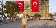 ein Mann steht ganz allein auf einem Platz. Im Hintergrund ein monumentaler Betonbau mit türkischen Fahnen. Die Sonne scheint