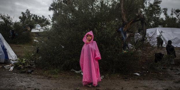 Ein Kind steht vor einem Baum in einem Flüchtlingslager, es trägt ein rosa Cape