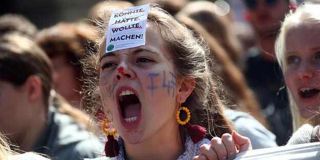 Eine junge Frau ruft etwas, sie hat sich eine Blat Papier mit den Worten "Könnte,Hätte, Wollte - Machen" auf die Stirn geheftet